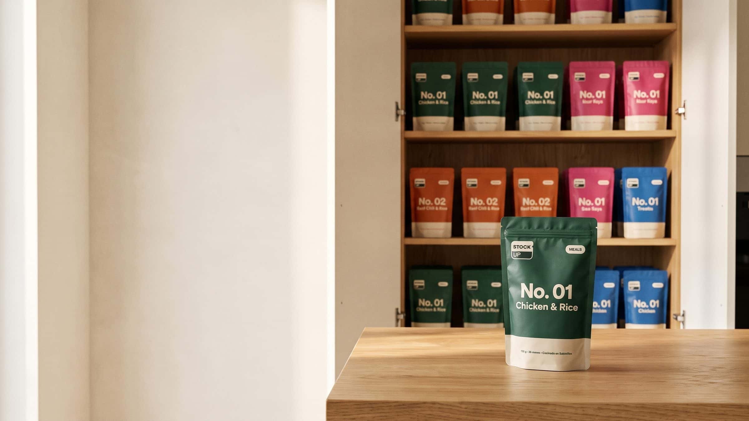 An open kitchen cupboard stocked with Stock Up pouches in all four colours, a green Meals No. 01 pouch on the worktop