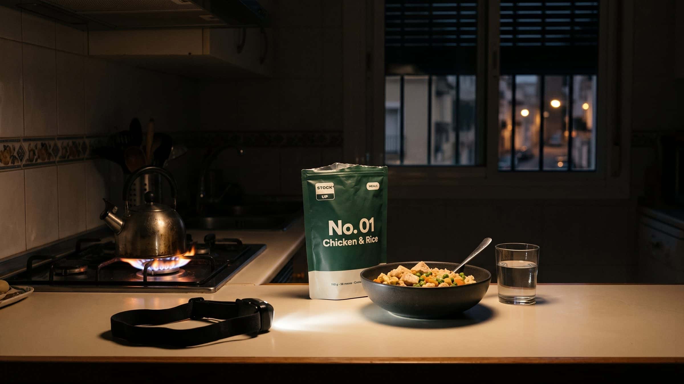 Cocina a oscuras en un apagón: fogón de gas encendido, un frontal, la bolsa Meals verde y un bol de pollo con arroz